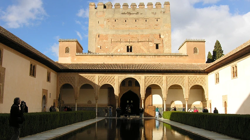 İspanya'nın Granada şehrindeki tarihi Alhambra Sarayı'nın yemyeşil çitlerle çevrili uzun yansıtma havuzunun iki yanında zarif kemerli revakların uzandığı, detaylı süslemelere sahip duvarların ve arka planda yükselen crenelli bir tuğla kulenin göründüğü, ziyaretçilerin dolaştığı geniş ve etkileyici bir avlu manzarası.