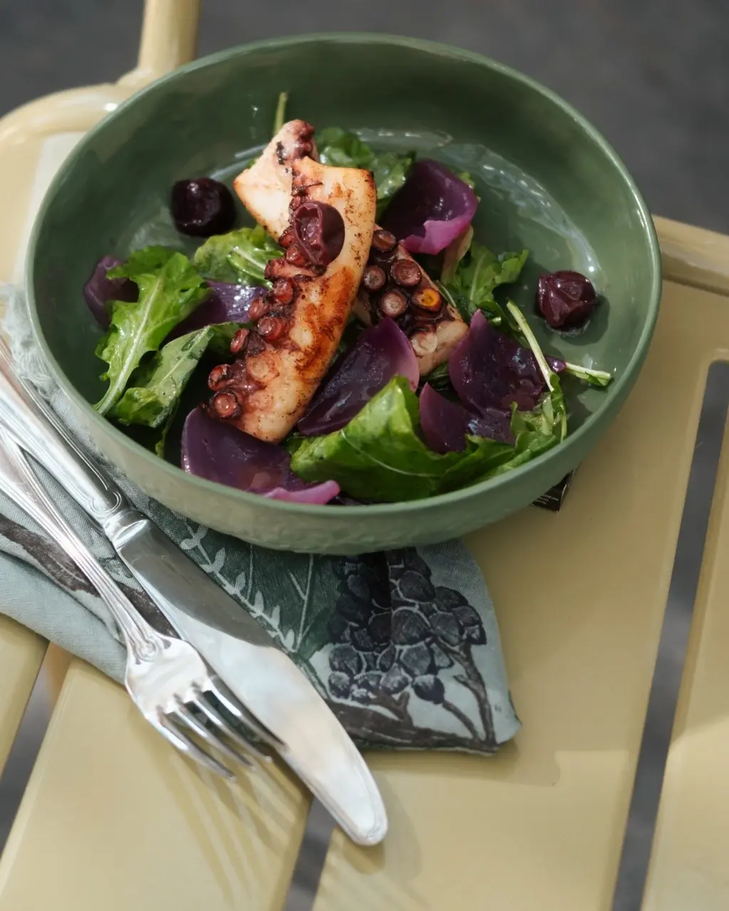 Urla'nın zengin gastronomisine özgü, ızgara ahtapot, taze yeşillikler ve mor renkli sebzelerle sunulan bu tabak, üzüm salkımı motifli bir peçetenin üzerinde yer alarak bölgenin denizden gelen lezzetleri ve Bağ Rotaları çevresindeki doğal ürünleriyle harmanlanmış güzelliğini vurguluyor.