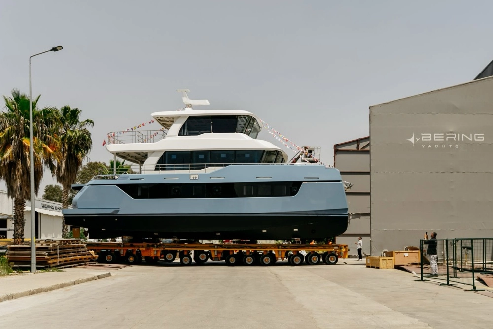 Açık alanda, Bering Yachts logosunun yer aldığı bir yapının yanında, çok tekerlekli bir taşıyıcı üzerinde taşınan, fildişi beyazı üst güverteye, gri-mavi renkte ana gövdeye ve lacivert alt gövdeye sahip, bayraklarla süslenmiş büyük bir Türk yapımı Bering BC60 katamaranı görülüyor.