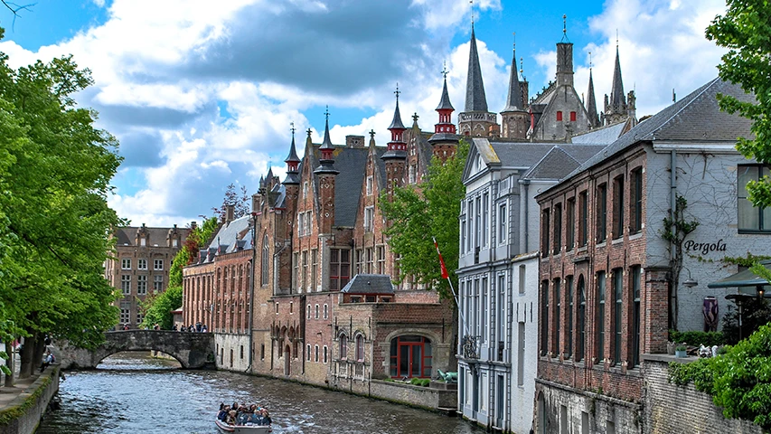 Belçika'nın tarihi Brugge şehrinde, bir köprü altından geçen, gezinti tekneleriyle dolu sakin bir kanal, her iki yanında Gotik mimarinin özelliklerini taşıyan detaylı çatılı ve kuleli eski tuğla binalarla çevrili, yeşillikler içindeki büyüleyici bir manzara sunuyor.