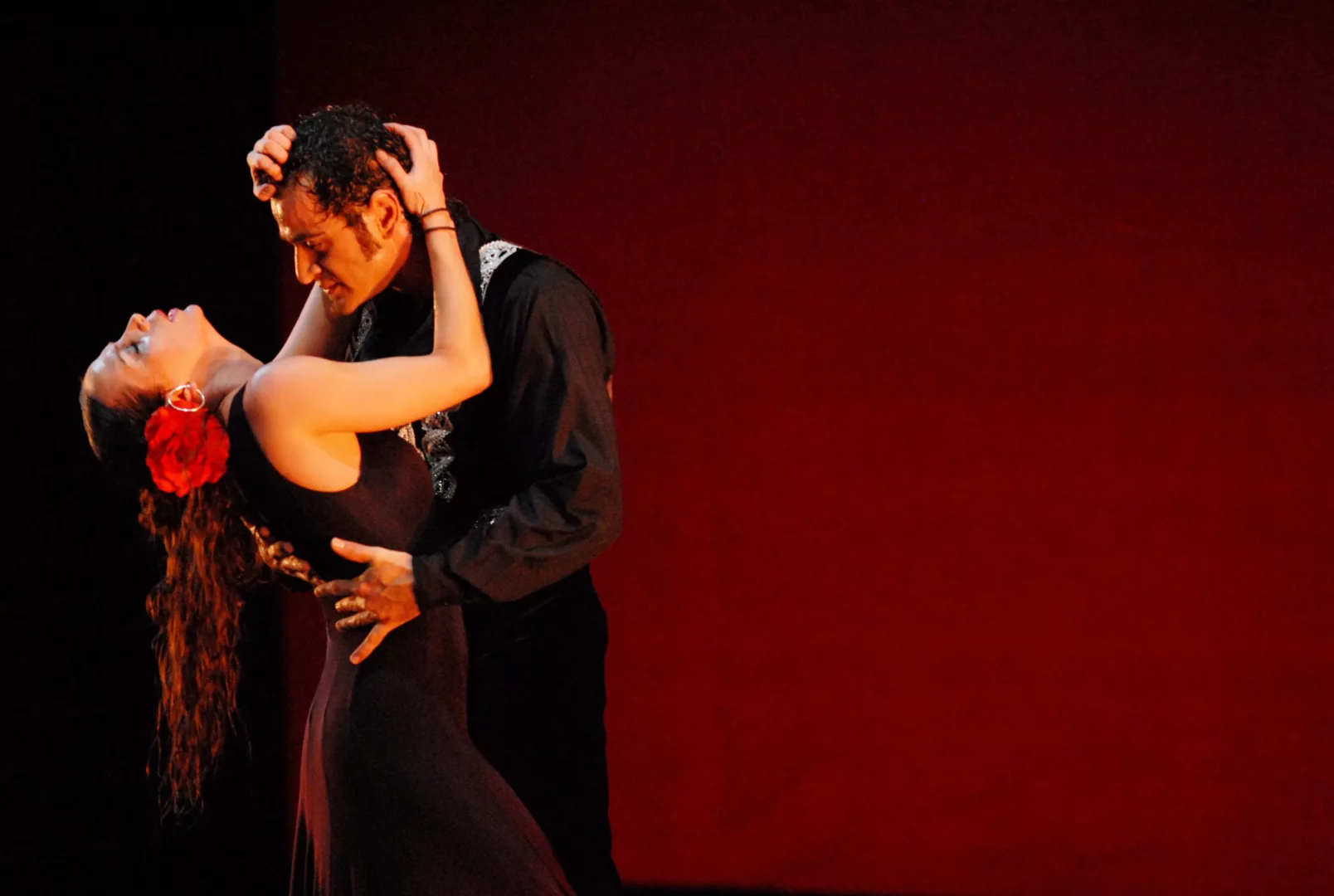 Koyu kırmızı bir sahne zemininde, Bizet’nin Carmen operasının tutkulu atmosferini yansıtan bir an: Siyah elbisesinin omuzları açıkta, saçında parlak kırmızı bir çiçekle başını geriye atmış güzel bir kadın, işlemeli siyah gömlekli bir adamla kucak kucağa dramatik bir dans figürü sergileyerek aşkın ve trajedinin derinliğini gözler önüne seriyor.