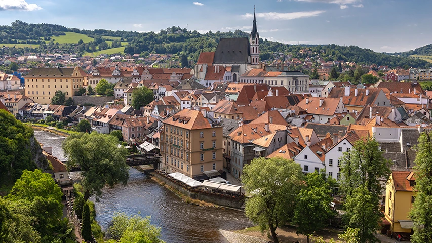 Çekya'nın tarihi Český Krumlov şehrinin, Vltava Nehri'nin kıvrımları boyunca sıralanmış, belirgin sivri kulesi olan Aziz Vitus Kilisesi ve kendine özgü kırmızı kiremitli çatılarıyla dikkat çeken binalarının yemyeşil tepelerle çevrili, güneşli bir günde çekilmiş geniş açılı panoramik görüntüsü.