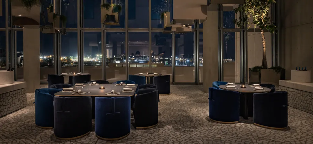 Geniş pencerelerinden gece ışıklarıyla parlayan bir şehir ve marina manzarası görünen, koyu mavi kadife sandalyelerle donatılmış şık masaların yer aldığı modern ve lüks bir restoran iç mekanı.