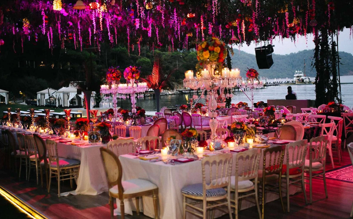 AC Design and Event Co. tarafından özenle tasarlanmış bu görüntüde, mor ve pembe tonlardaki ışıklandırmanın tavandan sarkan çiçeklerle bütünleştiği, deniz kenarında kurulan uzun yemek masalarının sayısız mum, kristal şamdanlar ve canlı çiçek aranjmanlarıyla bezendiği, rüya gibi bir düğün organizasyonu atmosferi yansıtılıyor.