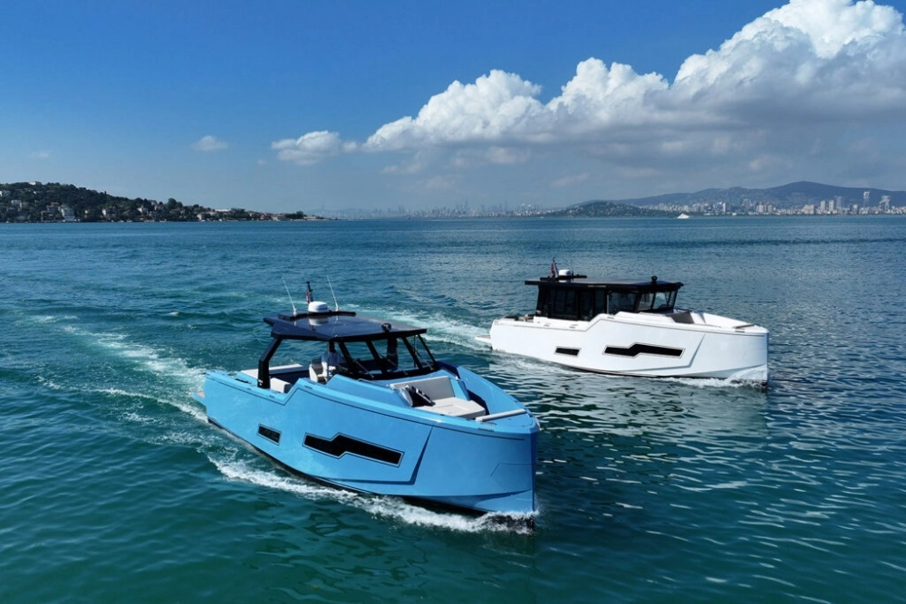 Parlak mavi renkli, ön tarafı açık bir Fabbro F45 coupe-cabrio tekne, turkuaz renkteki sakin denizde arkasında köpüklü bir iz bırakarak ilerlerken, sağ tarafında benzer modern hatlara sahip beyaz bir tekne de denizin tadını çıkarıyor, fonunda yemyeşil tepeler ve uzak bir şehir silüetiyle bulutlu masmavi bir gökyüzü dikkat çekiyor.