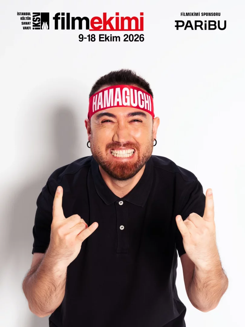 Kırmızı HAMAGUCHI yazılı saç bandı takan, sakallı, siyah polo tişörtlü, gözleri kısık ve neşeli bir ifadeyle dişlerini göstererek gülen adam, iki elini de rock on işareti yaparak Filmekimi festivalinin coşkulu ruhunu yansıtıyor.