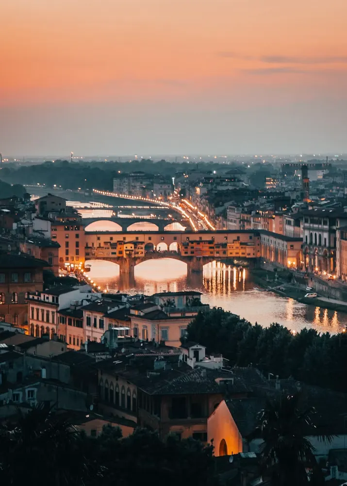 Alacakaranlıkta çekilmiş bu panoramik Floransa şehir manzarasında, Arno Nehri üzerinde ışıkları yansıyan kemerli köprüler, özellikle üzerinde binalar bulunan ikonik Ponte Vecchio'nun ışıltısı ve çevresindeki tarihi yapıların pencere ışıkları, şehrin canlı atmosferini sıcak turuncu ve pembe tonlara bürünmüş gökyüzünün altında vurguluyor.