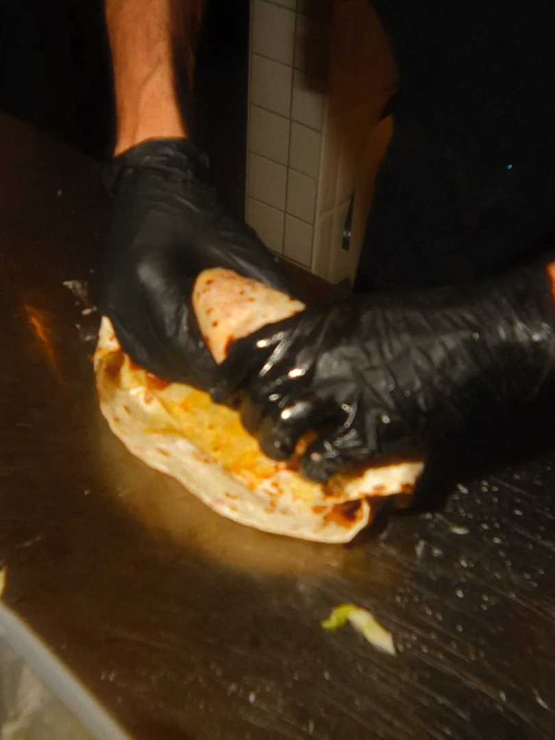 Siyah eldivenli eller, parlak metal bir tezgah üzerinde, içerisinde turuncu-sarı dolgu ve soslar bulunan büyük bir lavaşı dikkatlice sararak, Gringo Vegan mekanının lezzetli ve taze bir vegan dürümlerinden birini hazırlıyor.
