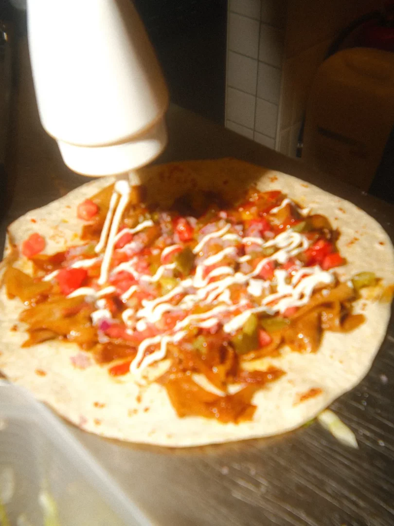 Geniş bir lavaşın üzerine bolca yerleştirilmiş kahverengi renkli vegan döner parçaları, kırmızı küp doğranmış domatesler, yeşil biberler ve mor soğanlar gibi çeşitli sebzelerle zenginleştirilmiş dolgunun üzerine, beyaz bir sos şişesinden kremsi beyaz mayonez özenle sıkılırken, Gringo Vegan'ın Moda'daki lezzetli vegan yemek hazırlık sürecini gösteren yakın çekim bir görüntü.