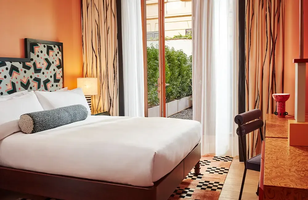 Turuncu duvarlara ve Memphis tarzı desenli yatak başına sahip modern bir otel odasında, beyaz çarşaflı yatağın hemen yanında yeşil bitkilerle dolu bir terasa açılan cam kapılar ve sanat eserini andıran özgün bir masa lambası görülüyor, bu şık tasarım alanı konfor ve estetiği bir araya getiriyor.