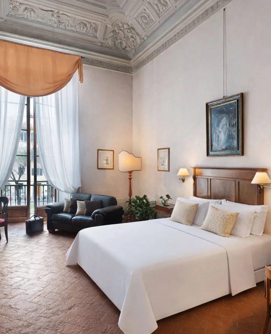 Palazzo Guadagni Oteli'nin, beyaz çarşaflı geniş yatağı, ahşap yatak başı, koyu renk deri kanepesi ve zarif işlemeli tavanıyla klasik bir ambiyans sunan odasında, geniş balkonlu pencerelerden dışarıdaki şehir manzarası görülmektedir.