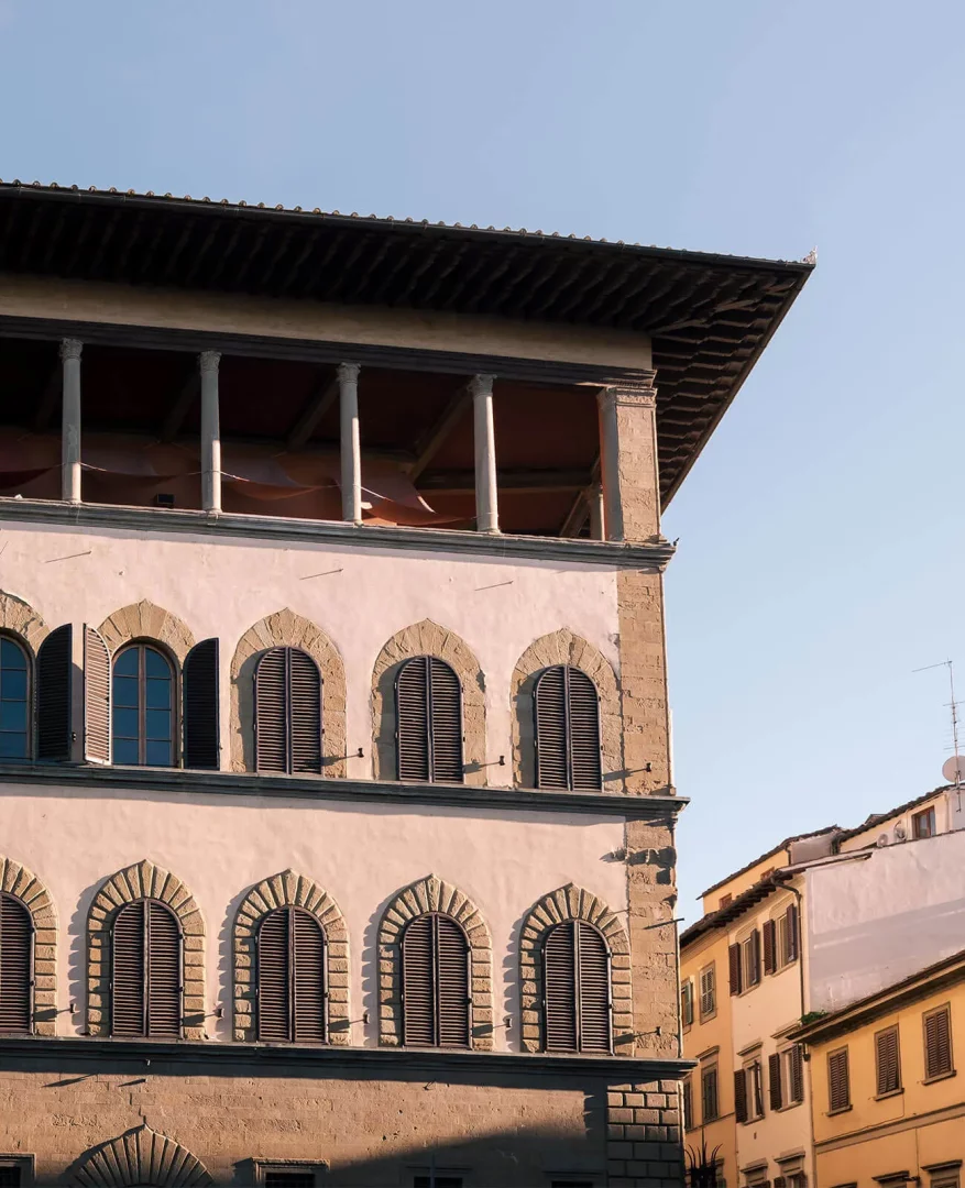 Hotel Palazzo Guadagni'nin kemerli pencereli, panjurlu cephesini ve en üst kattaki sütunlu loggia alanını yakalayan bu görsel, güneşte ışıldayan tarihi İtalyan mimarisinin zengin detaylarını sunuyor.