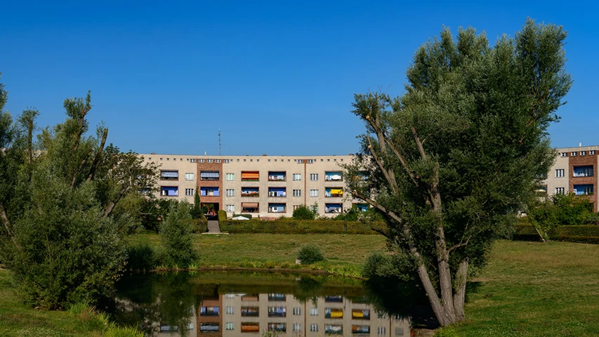 Masmavi bir gökyüzü altında, yemyeşil ağaçlar ve yansıtan bir göletle çevrili, açık renkli cephesi ve renkli pencereleriyle dikkat çeken çok katlı Hufeisensiedlung apartmanları görünüyor.