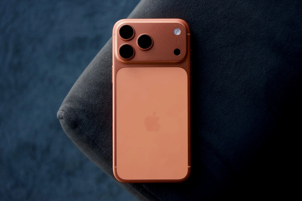 iPhone 17 Pro Max modelinin turuncu arka yüzü, koyu mavi/gri kumaş bir zeminde dikey olarak dururken, sol üst köşesindeki üçlü kamera lensleri, flaş ve sensör içeren çıkıntılı modülü ile ortasındaki Apple logosu ayrıntılı bir şekilde gösteriliyor.
