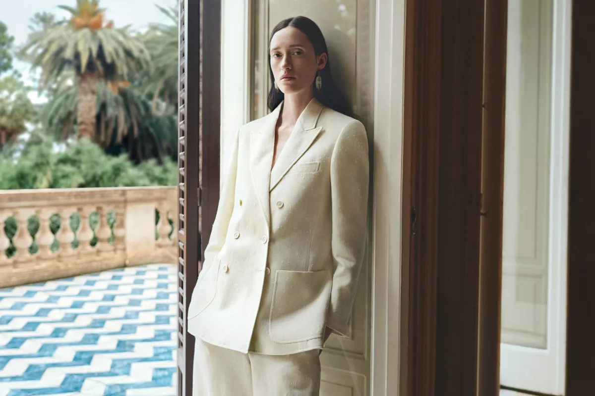 Kapı pervazına yaslanmış, üzerinde bej tonlarında, kruvaze ve oversize bir blazer ile uyumlu pantolon giymiş, uzun saçlı bir kadın, blazer'ların şık ve çok yönlü kullanımının altını çizen bu makale için ideal bir moda ilhamı sunuyor; arka planda palmiye ağaçları ve desenli karolarla Akdeniz esintileri taşıyan zarif bir dış mekan görülüyor.