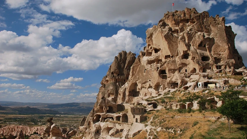 Kapadokya'da, beyaz bulutların süslediği masmavi gökyüzü altında, zirvesinde Türk bayrağının gururla dalgalandığı, tarih boyunca oyularak yaşam alanlarına dönüştürülmüş devasa Uçhisar Kalesi'nin ve uzanan vadinin büyüleyici doğal manzarası.