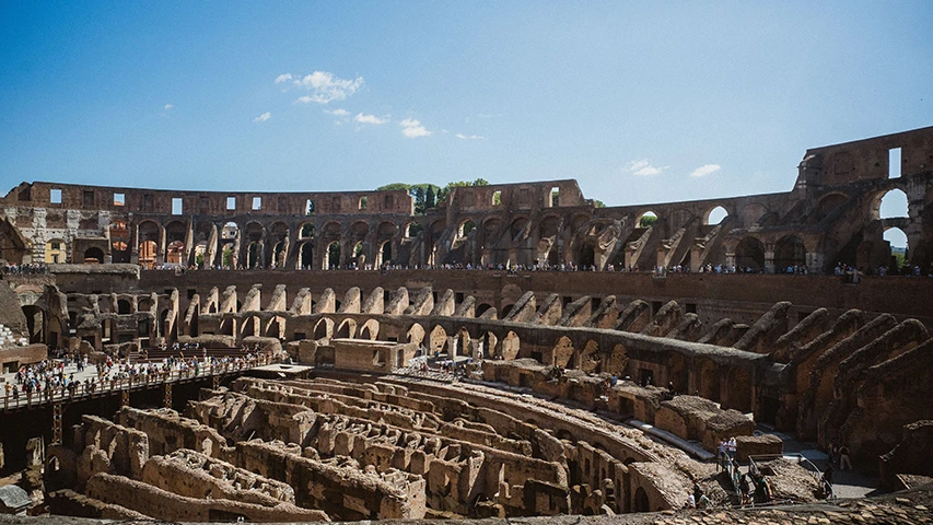 Roma'daki Kolezyum'un içinden çekilmiş geniş açılı bir fotoğrafta, yarı dairesel, taş yapıdaki antik amfitiyatronun yıkık arena katı ve seyirci basamakları tüm ihtişamıyla görülüyor; zemin katında karmaşık yeraltı odaları ve tüneller belirgin, üst katlarda ise kemerli açıklıklar ve dikdörtgen pencereler mevcut; güneşli havada yapıyı keşfeden çok sayıda turist, ahşap yürüme yollarında ve tarihi basamaklarda dolaşıyor.