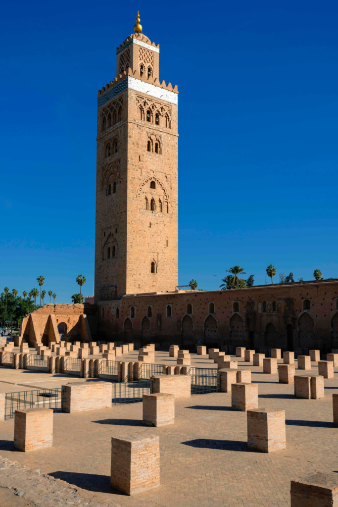 Marakeş'in ikonik minaresi Kutubiye Camii'nin zarif kiremit dokulu kulesi, mavi gökyüzü altında parıldayan altın alemi ve alt kısmındaki alçak tuğla sütunlarla çevrili geniş avlusuyla Marakeş Seyahat Rehberi'nde şehrin tarihi renklerini yansıtıyor.