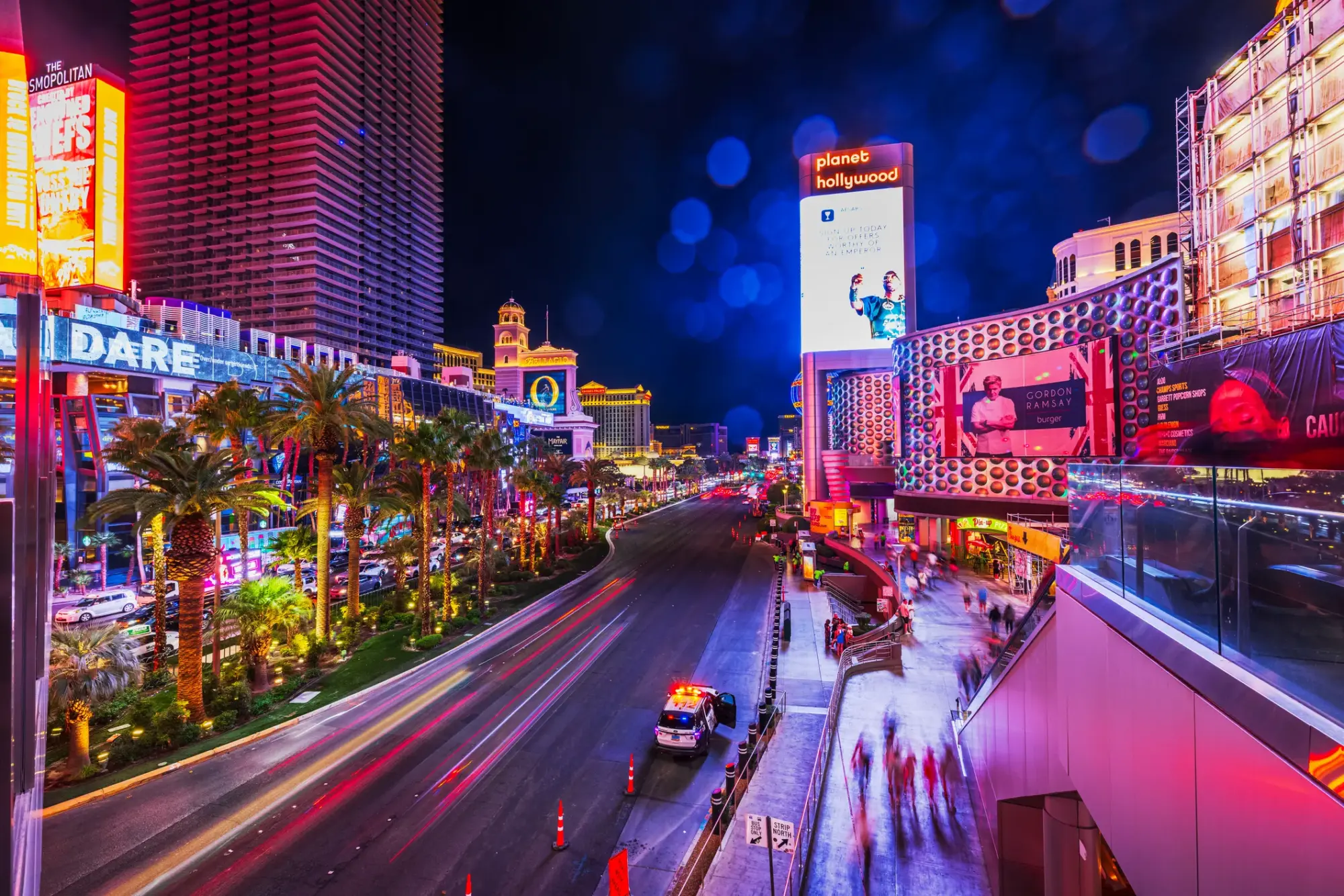 Las Vegas'ın meşhur Strip Caddesi'nin, Cosmopolitan ve Planet Hollywood gibi otellerin neon ışıklarıyla aydınlandığı, araç ışıklarının uzun pozlama efektiyle yol boyunca renkli çizgiler oluşturduğu, palmiyelerle çevrili bu hareketli gece manzarası, özel bir evlilik teklifi için büyüleyici ve unutulmaz bir fon sunuyor.
