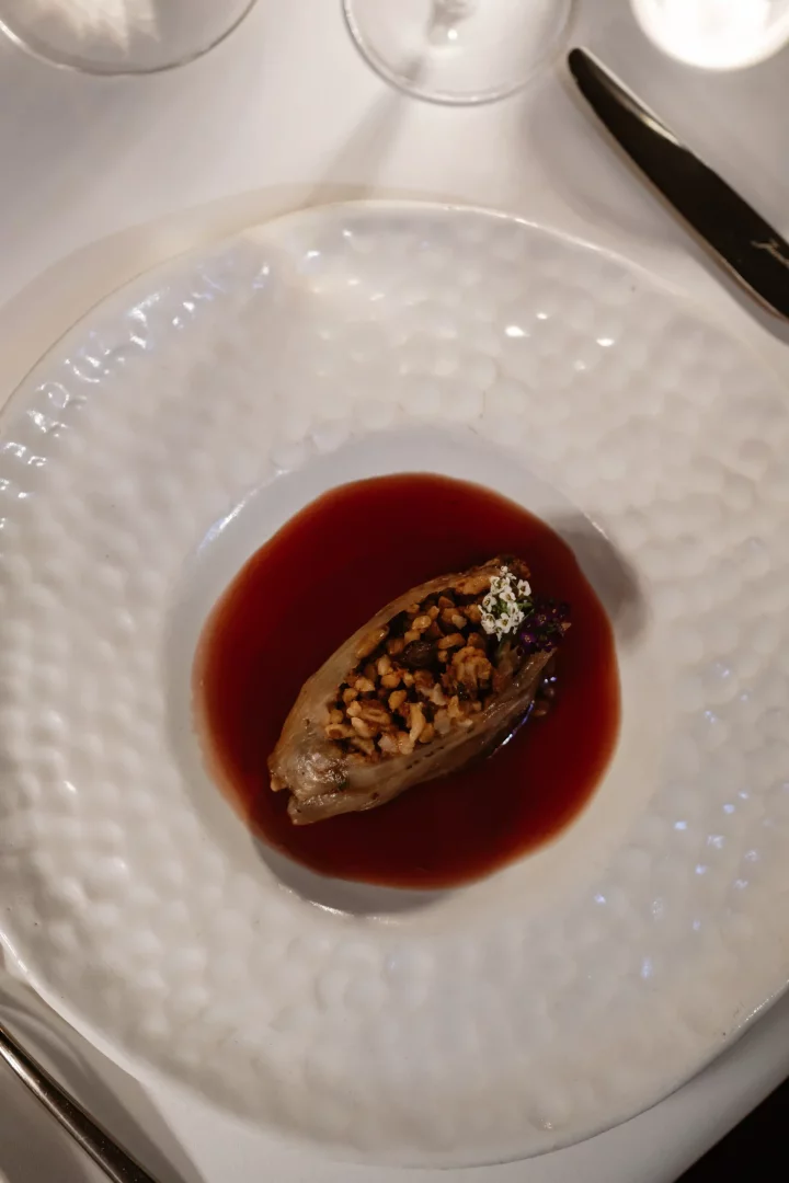 Lila Restaurant'ın sofistike mutfağından bir örnek olan bu görselde, pirinç ve kuruyemiş dolgulu, koyu kırmızı bir sosun içinde yüzen, zarif beyaz, dokulu bir tabakta sunulan, üstü narin beyaz ve mor çiçeklerle bezenmiş benzersiz bir lezzet vurgulanıyor.