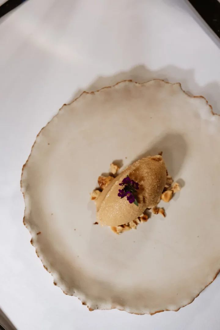 Museum Hotel'in Lila Restaurant'ında sunulan, üstünde minik mor çiçekler ve çevresinde kavrulmuş fındık parçacıklarıyla süslenmiş, açık kahve renkli bir porsiyon dondurma veya sorbe, düzensiz dalgalı kenarlara sahip, açık bej renkli seramik bir tabakta zarifçe duruyor.