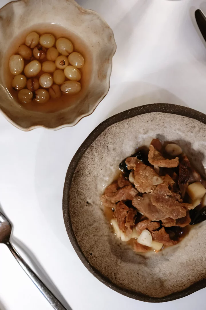 Lila Restaurant'ta, özenle hazırlanmış bir masada, açık renkli dalgalı seramik kasede tatlı şerbet içindeki zeytin veya üzüm taneleri ile rustik koyu renkli seramik kasede sarımsak ve mantar ile harmanlanmış kızarmış et parçalarından oluşan iki özel lezzet sunuluyor.