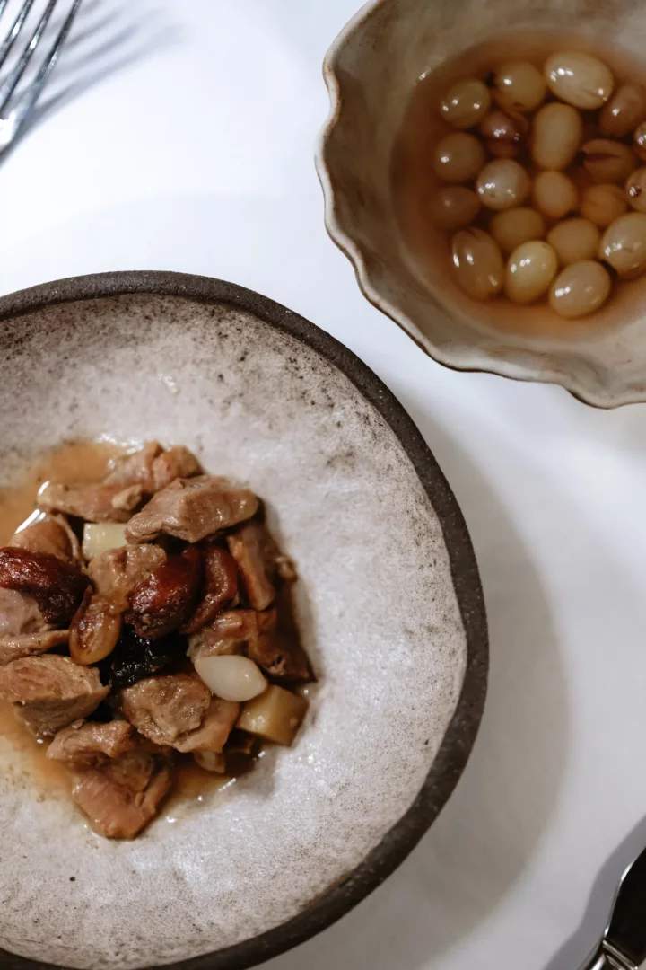 Lila Restaurant'ın özel menüsünden, rustik dokulu bir seramik kasede sunulan, koyu soslu, kuru meyveli ve minik patates veya sarımsak parçacıklı et yemeği, yanında dalgalı kenarlı küçük bir kapta hafif şerbet içinde yüzen açık renkli üzüm taneleriyle tatlı bir komposto ikramıyla şık bir masada yer alıyor.