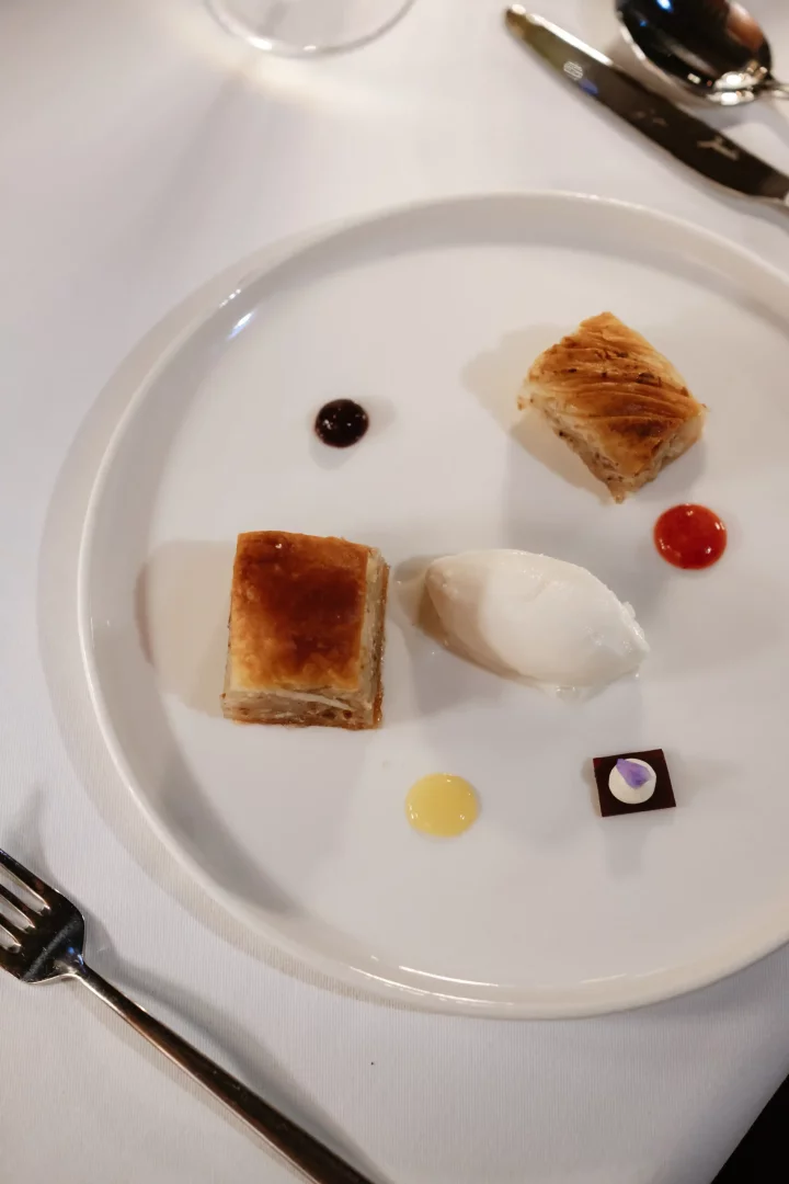 Beyaz bir yemek tabağında özenle yerleştirilmiş, hafifçe kızarmış iki dilim baklava benzeri tatlı, ortasında bir top beyaz dondurma, yanında farklı renklerde üç küçük sos damlası ve menekşe yaprağı süslü minik bir çikolata parçası, Museum Hotel bünyesindeki Lila Restaurant'ın özel tatlı sunumunu gözler önüne seriyor.