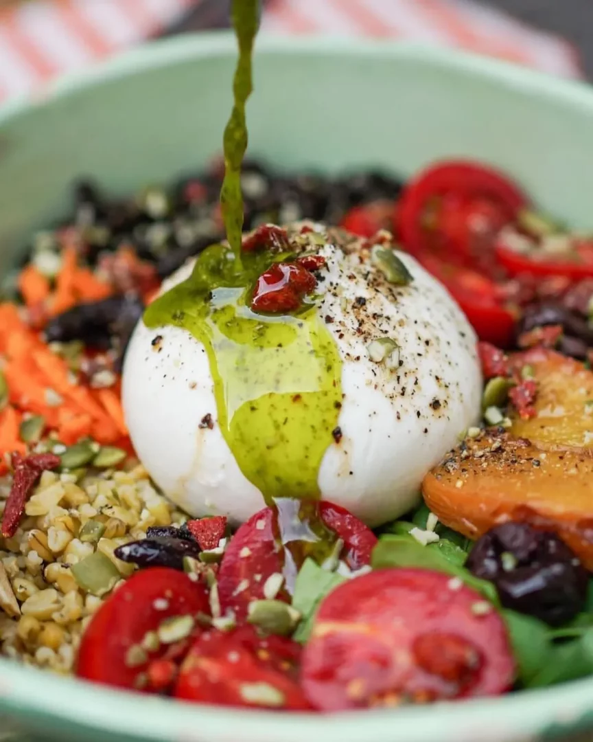 Yeşil bir kasede ortasında büyük, baharatlı bir burrata peyniri bulunan, üzerine taze yeşil pesto sosu akıtılan ve çevresinde taze dilimlenmiş kırmızı domatesler, kavrulmuş havuçlar, siyah zeytinler, balkabağı çekirdekleri ve bulgur pilavı ile zenginleştirilmiş iştah açıcı bir burrata pesto kasesi yemeği.