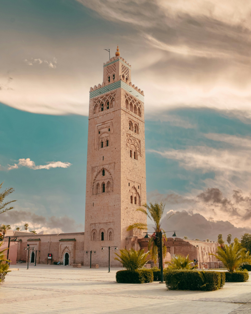 Marakeş'in ikonik Kutubiye Camii'nin zarif minaresi, oymalı taş işçiliği ve geometrik çini desenleriyle bezeli olarak, turkuaz ve sıcak tonlardaki bulutlu bir gökyüzünün altında, palmiye ağaçlarıyla çevrili ferah bir meydanda adeta renklerle uyanan bir rüya gibi yükseliyor.