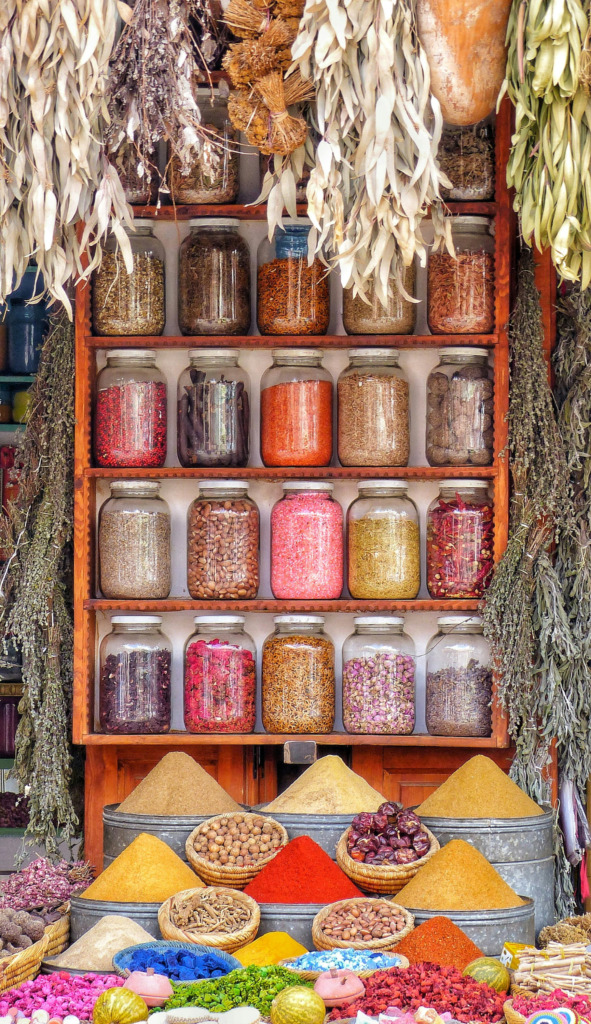 Marakeş'in canlı bir baharat pazarında, ahşap raflarda sıralı, farklı renklerde baharatlar, kurutulmuş güller ve otlarla dolu cam kavanozların altında, kırmızı, sarı, kahverengi gibi göz alıcı tonlarda konik baharat tepeleri ve hasır sepetlerde çeşitli ürünler sergileniyor, üst kısımdan sarkan kurutulmuş bitki demetleriyle Marakeş'in zengin renk ve baharat kültürü vurgulanıyor.