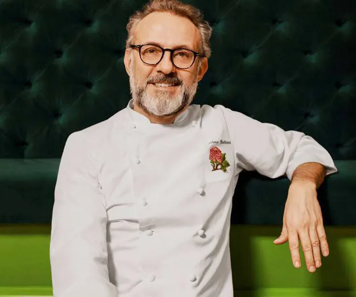 Ortadan hafifçe sağa dönük oturan, gözlüklü, gri sakallı şef Massimo Bottura, beyaz aşçı ceketinin sol omzunda zarif bir çiçek işlemeyle kameraya gülümserken koyu yeşil bir arka planın önünde poz veriyor.