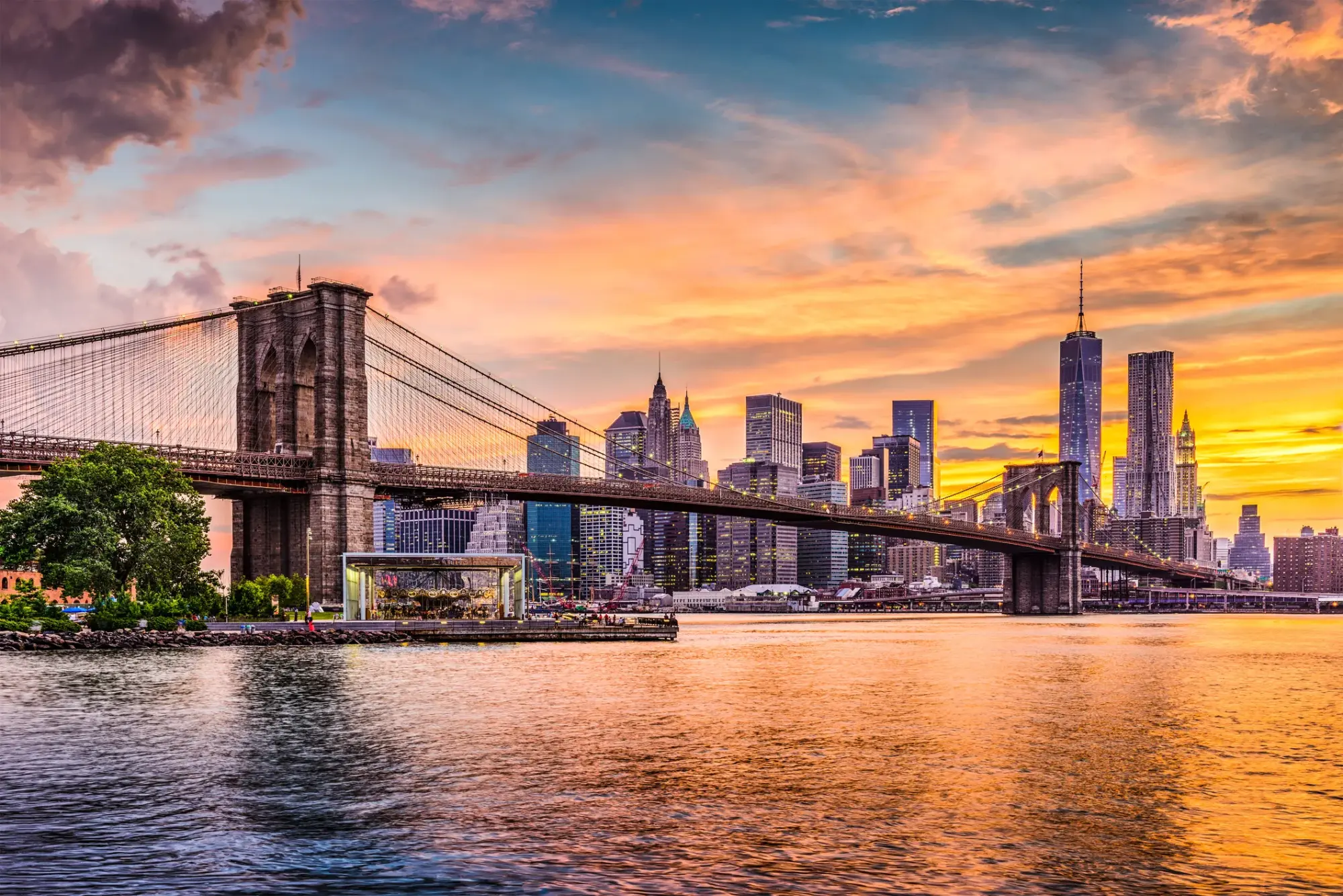 Gün batımının büyüleyici turuncu ve mor tonlarının gökyüzünü kapladığı bu geniş panoramik görünümde, New York'un meşhur Brooklyn Köprüsü, Manhattan'ın ışıklı gökdelenleri ve nehrin kıyısındaki Jane's Carousel ile birlikte eşsiz bir şehir manzarası sunuyor.