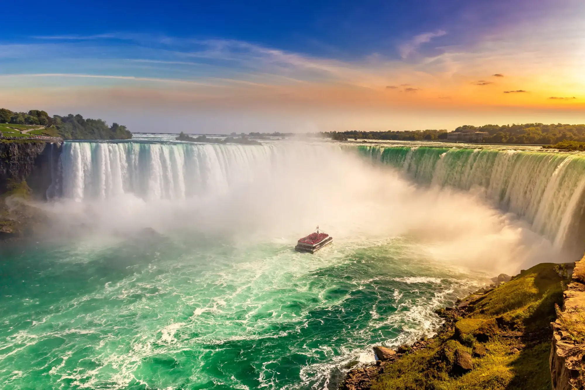 Güçlü Niagara Şelalesi'nin buharla kaplı coşkulu sularının ortasında, üzerindeki kırmızı pançolu turistlerle dolup taşan bir gezi teknesi ilerlerken, batmakta olan güneşin mavi ve turuncu tonlarındaki gökyüzüyle aydınlattığı görkemli bir doğa harikası.
