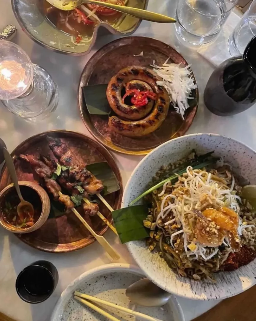 Nişantaşı'ndaki Nid Noi restoranında bir masanın üzerinde, ızgara sosis, şişte et ve karidesli Pad Thai gibi Tay mutfağı lezzetlerinin sunulduğu, mum ışığıyla aydınlatılmış keyifli bir akşam yemeği manzarası. İstanbul’daki Tay restoranları arasında yer alan Nid Noi Nişantaşı mekânından bir görünüm.