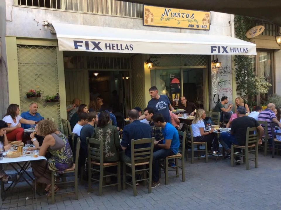 Beyaz bir tente üzerinde FIX HELLAS yazan, Nikitas adlı Yunan tavernasının önündeki kaldırımda, kalabalık bir grup insan yeşil sandalyelerde oturmuş, garsonun servis yaptığı masalarda sohbet edip yemek yiyerek keyifli bir açık hava mekanı atmosferi sunuyor.