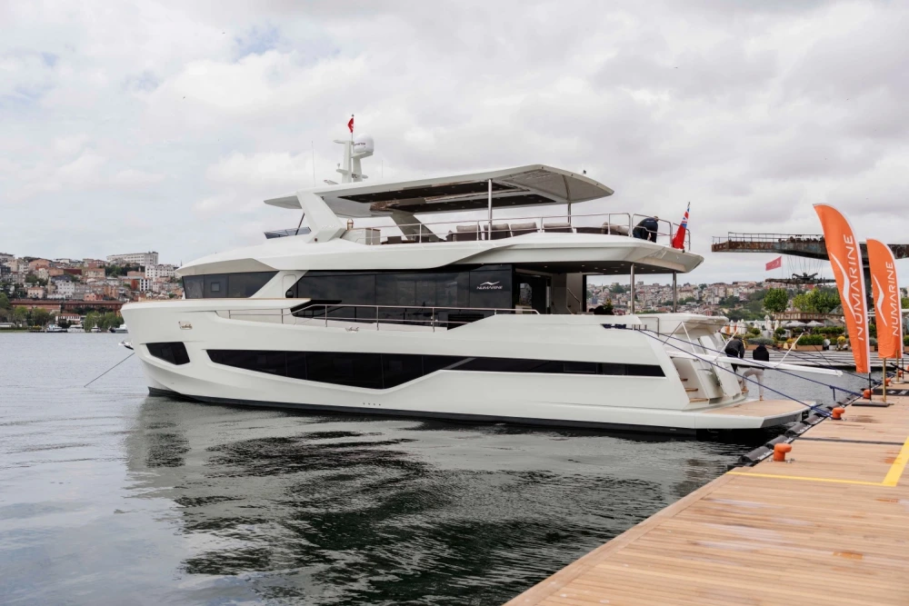 Beyaz ve modern çizgili Numarine 27XP explorer yatı, İstanbul'daki bir lansman etkinliği kapsamında ahşap iskeleye bağlı şekilde sakin sularda duruyor, arka planda şehrin silüeti ve bulutlu gökyüzüyle çevrili.