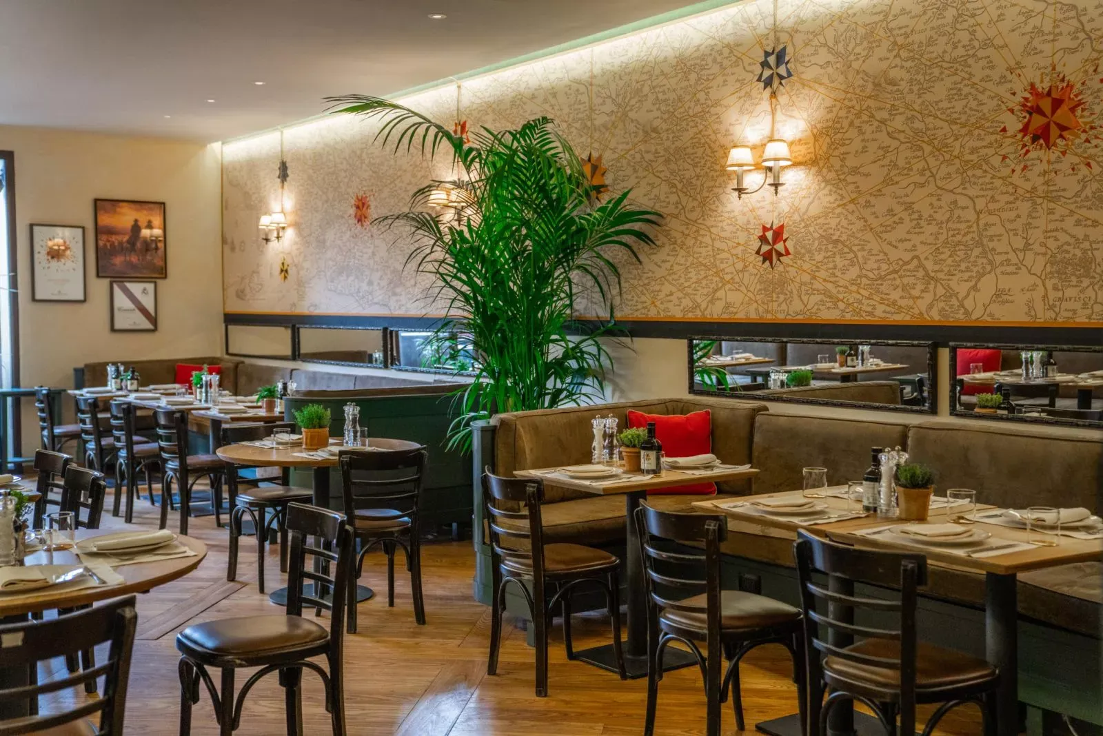 Haritalı duvar kağıdı ve pusula gülü süslemeleriyle dikkat çeken bir osteria veya İtalyan restoranının iç mekanı, şık ahşap masalar, rahat kahverengi banklar, kırmızı minderler ve ortamı canlandıran büyük bir palmiye ağacıyla misafirlerini ağırlamaya hazır, sıcak ve davetkar bir atmosfere sahip.