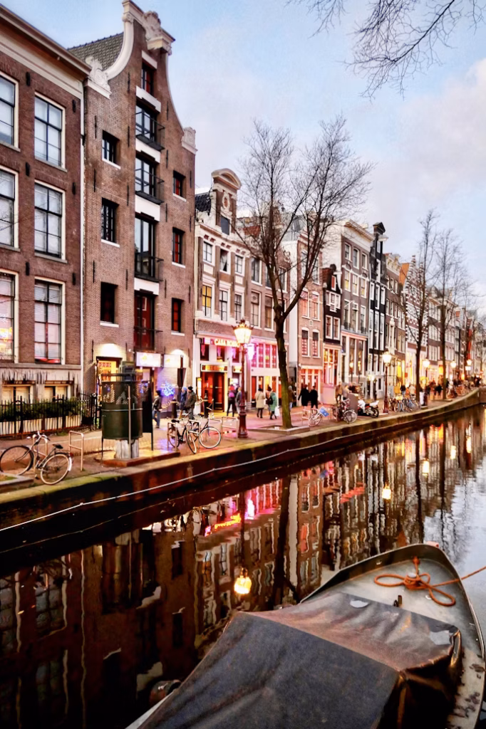 Gün batımının kızıllığı ve dükkan ışıklarının renkli yansımalarıyla aydınlanan Amsterdam'ın eşsiz kanallarından birinde, geleneksel dar cepheli tarihi evlerin ve kaldırımda yürüyen insanların olduğu hareketli bir akşam manzarası, ön planda bağlı bir tekneyle büyüleyici bir şekilde betimleniyor.