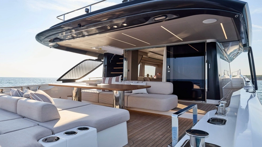 Prestige F6.7 flybridge yatın lüks kıç güvertesi, zarif bej minderli geniş oturma alanları, ahşap masa, üst güverteye giden modern merdiven ve masmavi denizin huzurlu manzarasıyla açık havada konforlu bir dinlenme ortamı sunuyor.