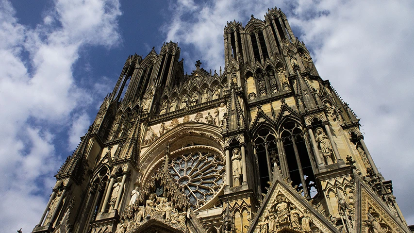 Alttan yukarıya doğru çekilmiş bu görkemli fotoğrafta, beyaz bulutlarla bezeli mavi bir gökyüzünün altında yükselen Reims Notre-Dame Katedrali'nin Gotik mimarisinin tüm detayları, zarif kemerleri, heykelleri ve tam ortasında yer alan etkileyici gül penceresi net bir şekilde görülüyor.