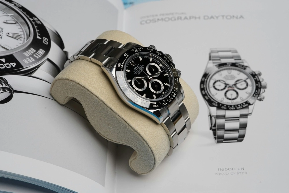 Bej renkli bir saat yastığı üzerinde parlak çelik bilezikli, siyah kadranlı ve belirgin kronograf alt kadranlarına sahip, şık bir Rolex Cosmograph Daytona erkek kol saati sergileniyor; arka planda ise aynı modelin farklı versiyonlarının bulunduğu bir katalog sayfası görünüyor.