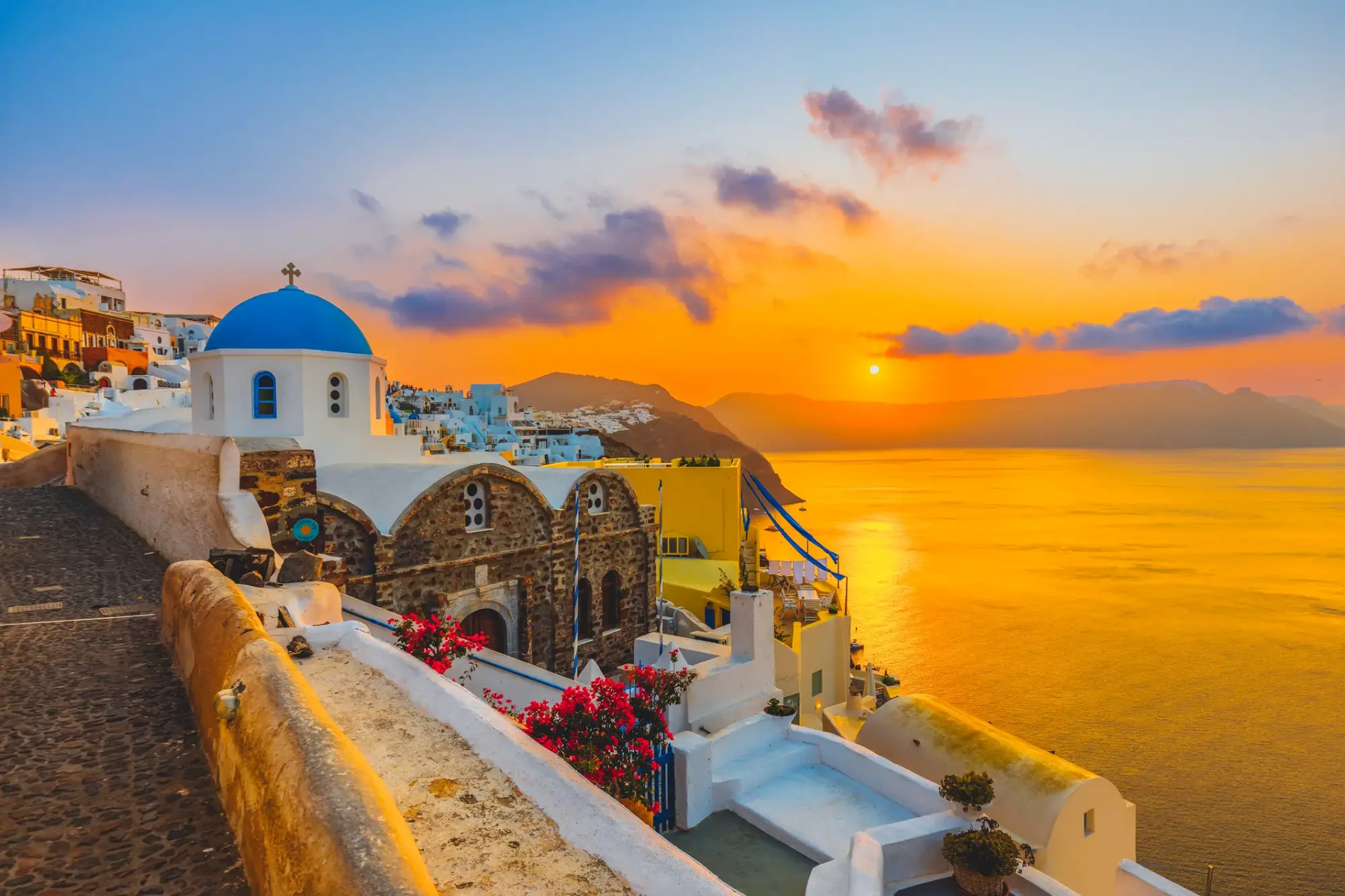 Santorini'nin Oia kasabasında, masmavi kubbeli ikonik bir kilisenin ve beyaz badanalı evlerin Akdeniz'in altın rengi gün batımıyla harmanlandığı, evlilik teklifi gibi özel anlar için unutulmaz bir fon oluşturan büyüleyici bir kaldera manzarası.