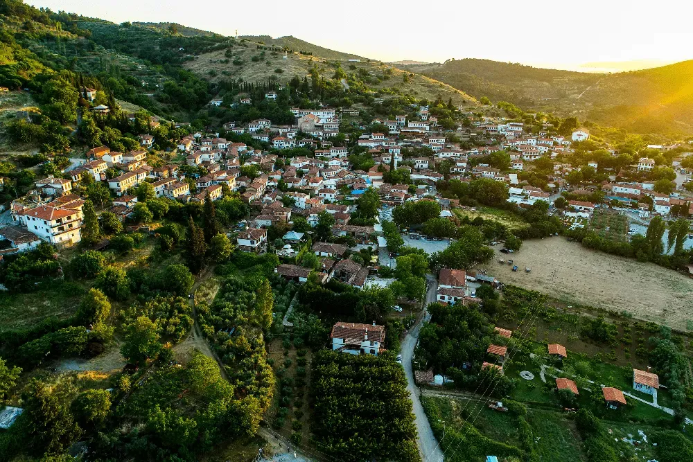 Gün batımının altın rengi ışıkları altında, yemyeşil tepelerin arasına serpilmiş, bembeyaz evleri ve kiremit çatılı geleneksel dokusuyla Şirince köyünün kuşbakışı muhteşem manzarası, bölgenin bağ rotalarına uygun doğal güzelliğini sergiliyor.