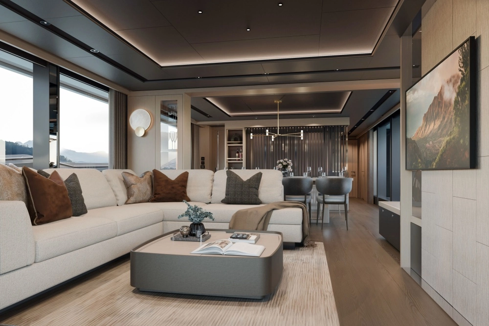 İngiliz yapımı lüks Sunseeker Manhattan 94 süper yatın aydınlık ve modern iç mekanında, büyük pencerelerden görünen dağ manzarası eşliğinde açık renk L şeklinde bir koltuk takımı, şık bir orta sehpa, televizyon ve zarif bir yemek alanı, kahverengi ve gri yastıklarla sıcak bir ambiyans sunuyor.