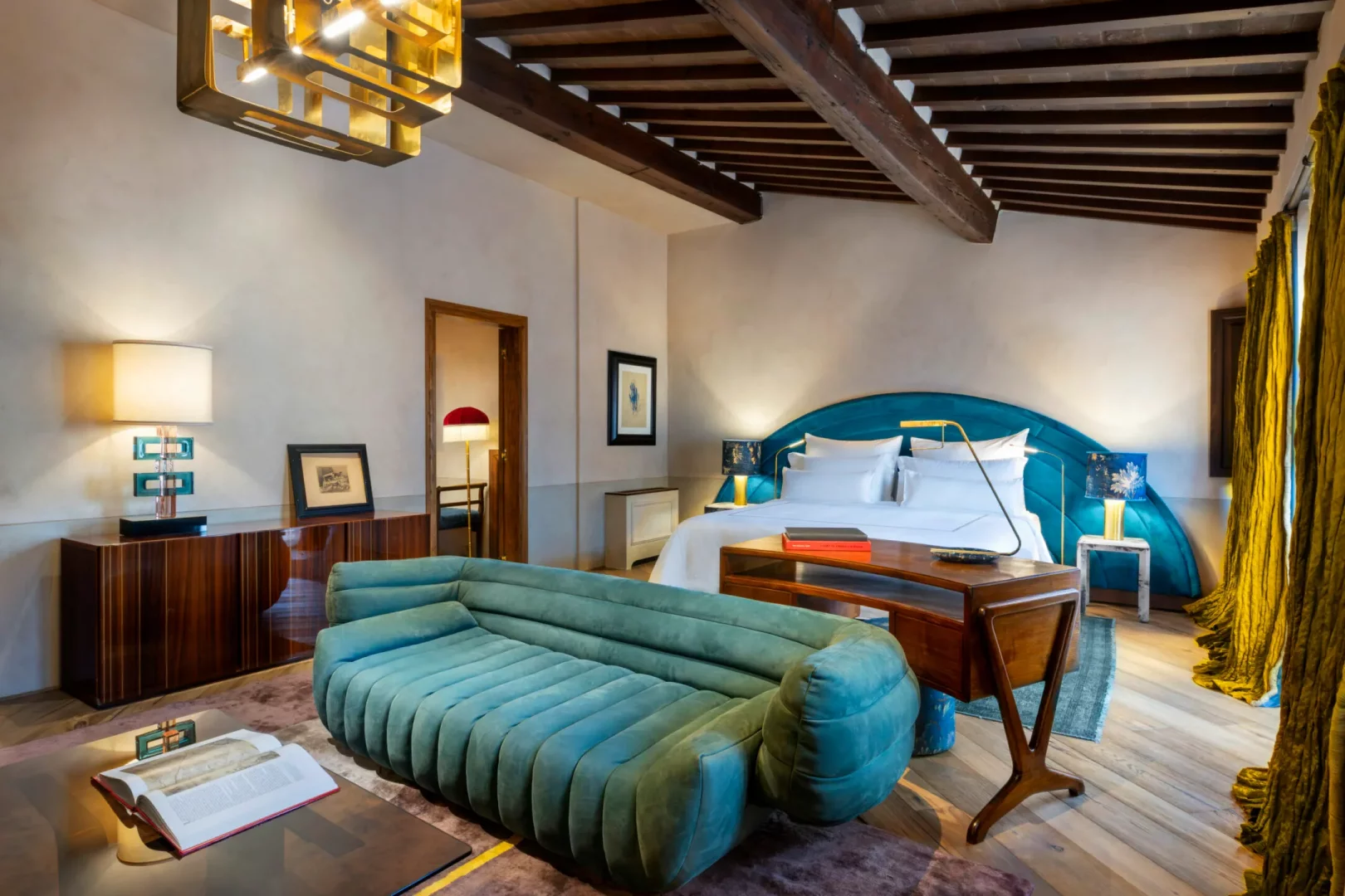 Lüks bir otel süitinde, kavisli teal kadife yatak başlığına sahip büyük yatak, önünde kanallı teal kanepe, zarif ahşap mobilyalar, altın renkli modern avize ve hardal sarısı perdelerle zenginleştirilmiş, tavanda görünen ahşap kirişlerin de eşlik ettiği şık bir iç mekan tasarımı.