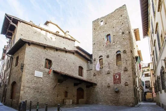 Floransa'da, pencerelerinde İtalyan bayrakları asılı, ön yüzünde Casa di Dante Müzesi'nin belirgin kırmızı tabelası bulunan yüksek taş kule ve ona bitişik tarihi taş binalarla çevrili, ortasında eski bir kuyunun olduğu dar bir meydanı gösteren geniş açı bir görüntü.