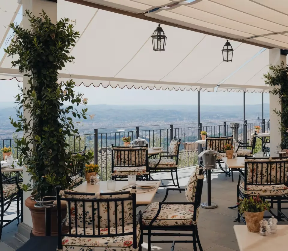 Villa Belmond'un açık hava yemek terası, beyaz tenteler altında düzenlenmiş çiçek desenli minderli sandalyeler ve masalarla, uzakta parlayan tepeler ve şehir silüetini gözler önüne seren nefes kesici bir manzaraya sahip.