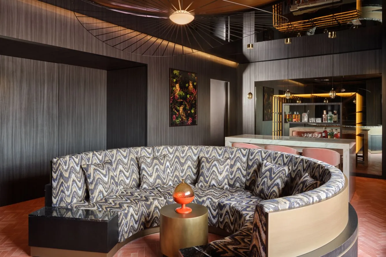 W Hotel'in modern tasarım anlayışını yansıtan bu lüks iç mekanda, lacivert ve krem rengi zikzak desenli, kavisli büyük bir koltuk takımı ortasında bronz bir sehpa ile birlikte dikkat çekiyor; koyu ahşap panelli duvarlar, tavan yansımaları ve arka plandaki şık bar alanı, odanın sofistike atmosferini tamamlıyor.