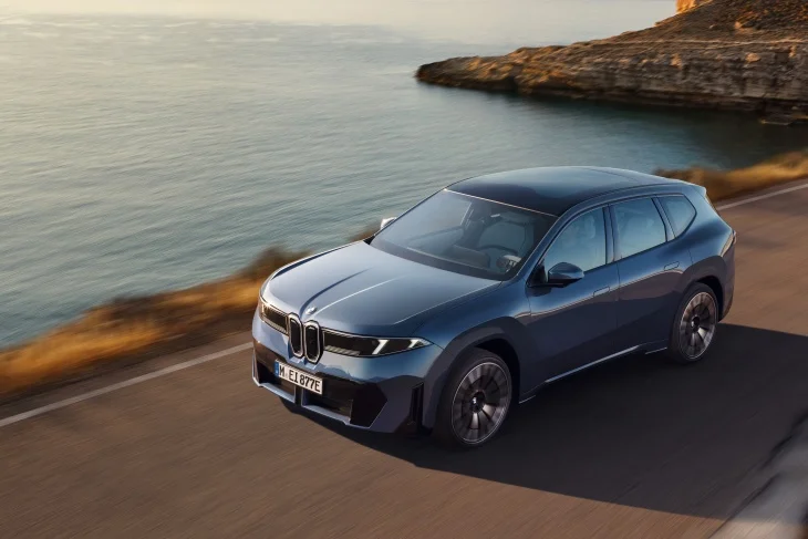 Kıyı şeridi boyunca uzanan bir yolda ilerleyen, koyu mavi renkli şık BMW iX3 elektrikli SUV, Neue Klasse tasarım dilinin sade ve modern çizgilerini sergileyerek markanın elektrikli geleceğini yollara taşıyor.